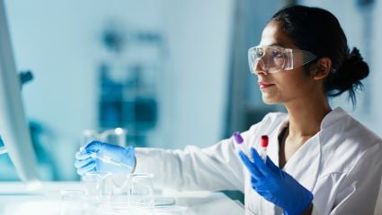 A female lab scientist with dark brown hair wearing safety goggles working with test tubes