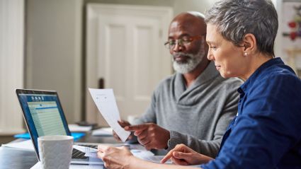 An elderly couple read paperwork in front of a laptop.