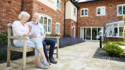 An elderly couple sits on a bench outside of a senior housing facility.