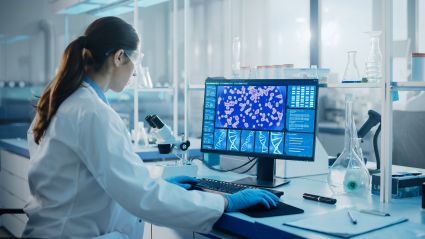 An image of a woman with white skin and brunette ponytail who is a scientist working in a lab. She is using a computer with technical items on the screen and wearing a white lab coat.