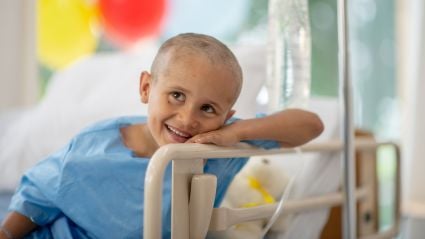 A mixed race oncology patient sits in his hospital bed, in his gown, with his hair shaved. He has an optimistic smile on his face as he receives his Chemotherapy via IV drip treatment.