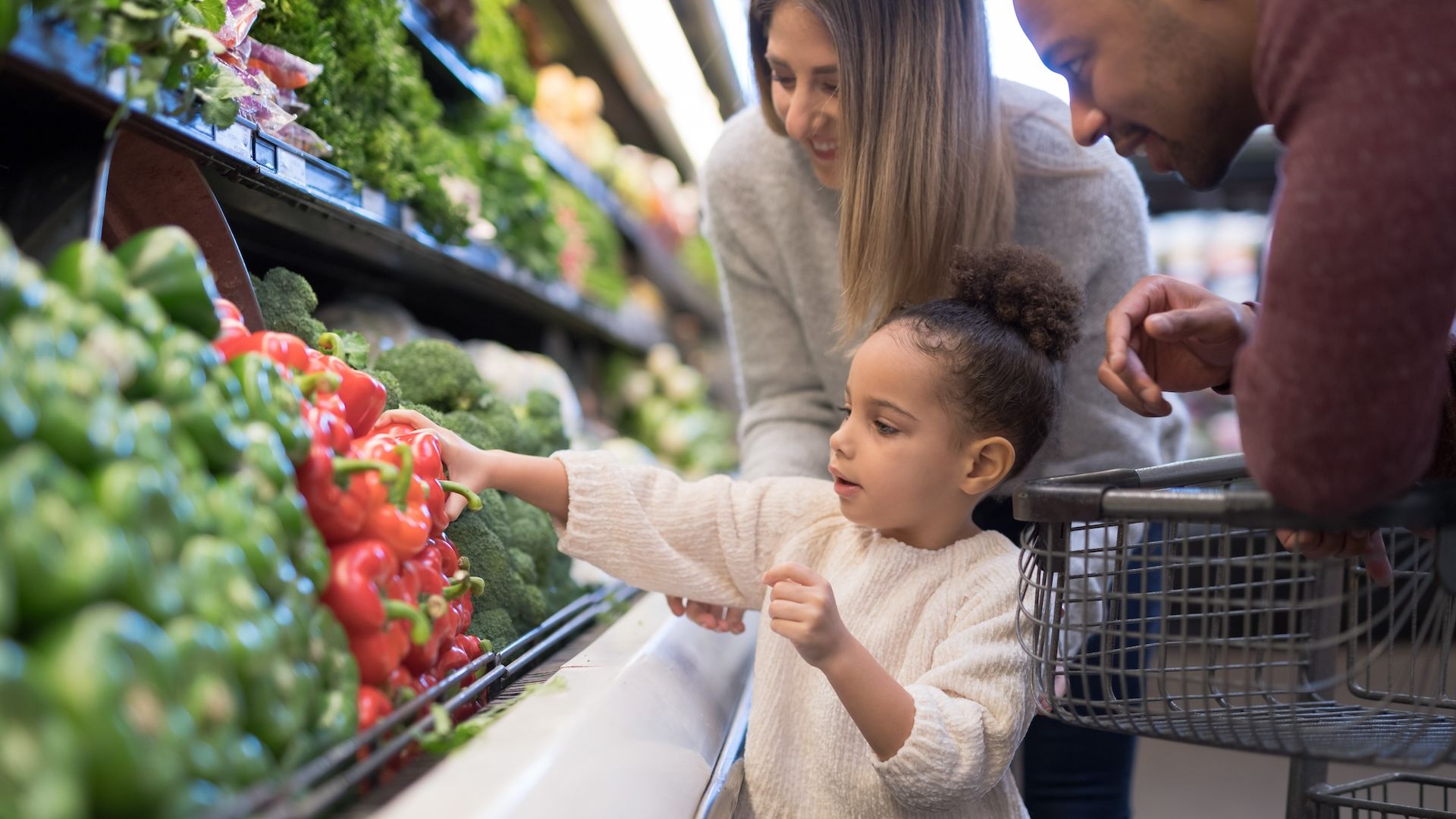 A male and female stand with young daughter in a grocery store aisle as she reaches for a red pepper in the produce section