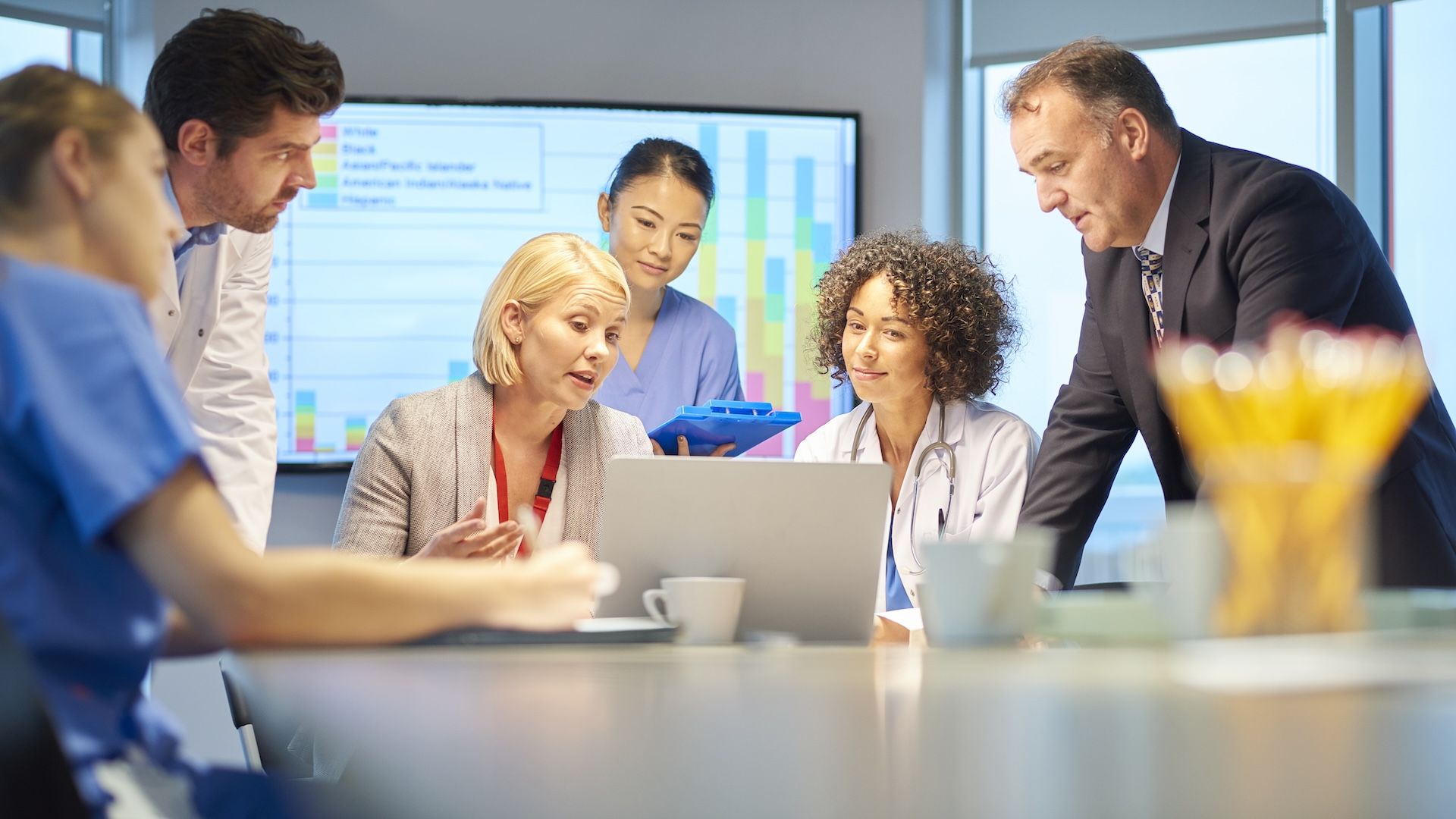 Six individuals sit around a conference table discussing something on a laptop screen in front of them