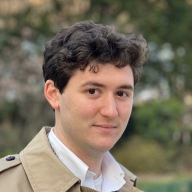 A light-skinned male-presenting individual with dark brown hair wearing a tan trench coat and white shirt against an outdoor backdrop