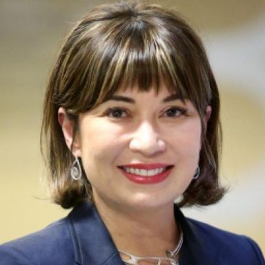 A female-presenting person with dark hair wearing a blue blazer against a light background.