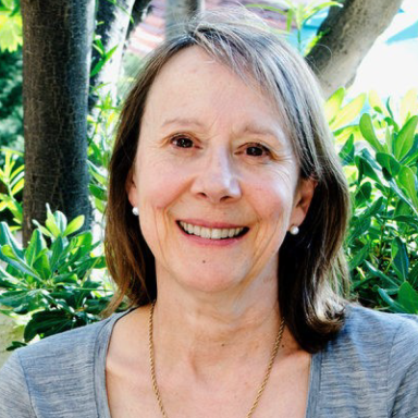 A smiling light-skinned female-presenting person with brown hair wearing a gray shirt against an outdoor backdrop