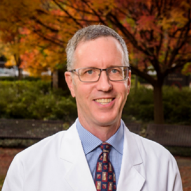 A smiling light-skinned male-presenting individual with gray hair wearing glasses, a white lab coat, a blue button-up shirt, and tie against an outdoor background