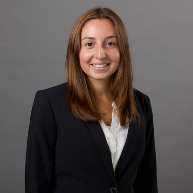 A smiling light-skinned female-presenting person with brown hair in a white shirt and dark suit jacket against a gray background.