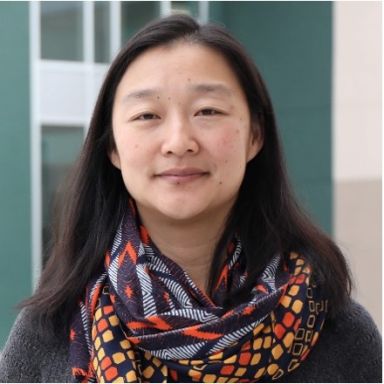 A female-presenting person with black hair wearing a dark top and a patterned scarf, against a blurred background.