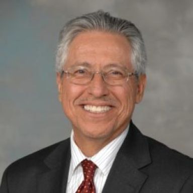 A smiling male-presenting person wearing eye glasses and a black blazer against a gray background.