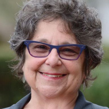 A smiling female-presenting person wearing eye glasses against a blurred background.