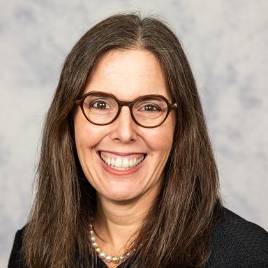 A smiling light-skinned female-presenting person with brown hair wearing a dark shirt against a grey background.