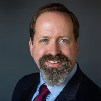 Smiling light-skinned, male-presenting individual with short dark hair wearing a dark suit on a grey backdrop.