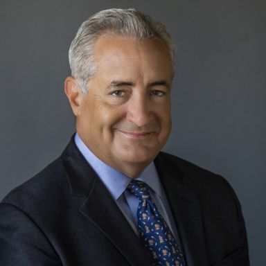 Smiling light-skinned, male-presenting individual with short grey hair wearing a dark suit on a grey backdrop.