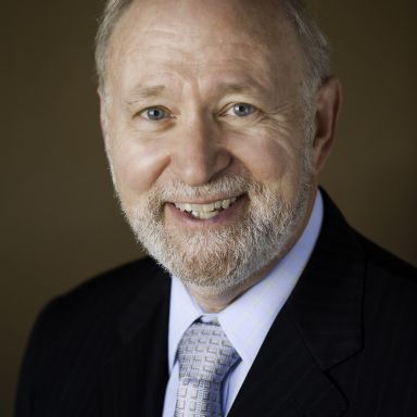 Smiling light-skinned, male-presenting individual with short grey hair wearing a dark suit on a brown backdrop.