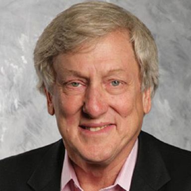 A smiling male-presenting person wearing a black blazer against a light gray background.