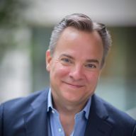 A light-skinned male-presenting person with short, dark hair wearing a suit jacket against a blurred  background