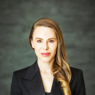 A light-skinned female-presenting person with long, red hair wearing a suit against a blurred dark background