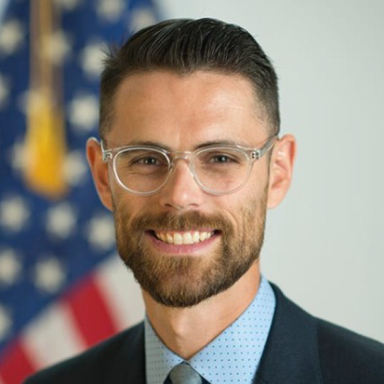 A smiling light-skinned, male-presenting individual with dark brown hair and a beard wearing glasses and a dark suit with a tie against a light background containing the American flag