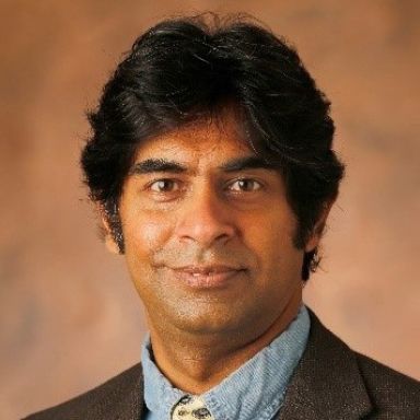 A darker-skinned, male-presenting person with black hair wearing a dark gray suit and tie against a brown gradient background