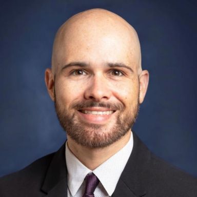 A smiling light-skinned male-presenting person wearing a white shirt with a dark tie and a black suit jacket against a navy blue background.