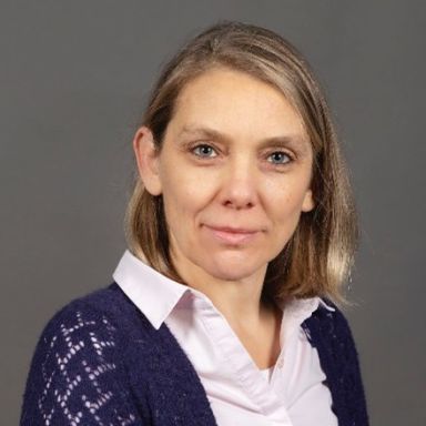 A light-skinned female-presenting person with light hair wearing a dark purple sweater over a button-up shirt in front of a dark gray background