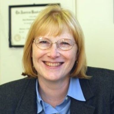 Smiling light-skinned female-presenting individual with light hair and glasses wearing a dark suit jacket with a blurred background