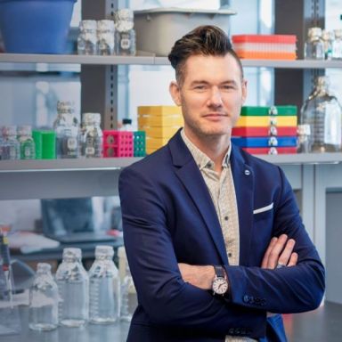 Light-skinned male-presenting individual with brown hair standing with arms crossed wearing a navy blue suit jacket and button-up against a set of shelves in the background