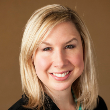 A smiling Caucasian female-presenting individual with blonde hair wearing a dark suit jacket against a brown gradient background