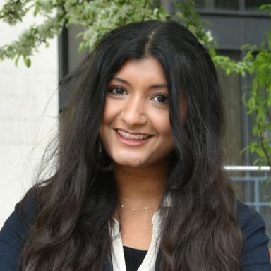 A smiling dark-skinned female-presenting person with dark hair against an outdoor backdrop.