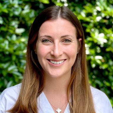 A smiling caucasian female-presenting person with brown hair wearing a white shirt against a green foliage background