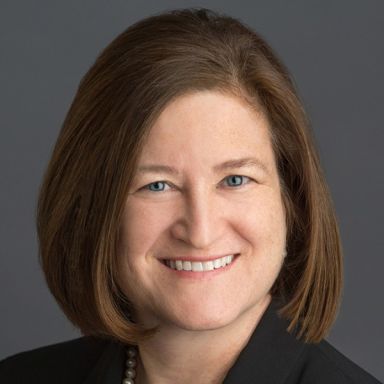 A smiling light-skinned, female-presenting person with dark brown hair wearing a dark suit jacket against a gray background