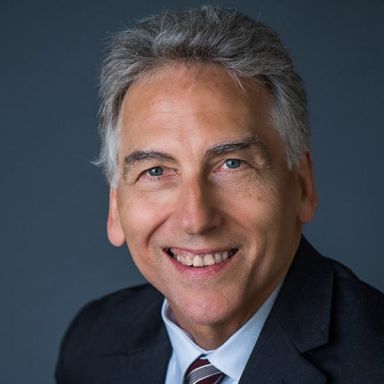 Smiling light-skinned, male-presenting individual with short grey hair wearing a dark suit on a grey backdrop.