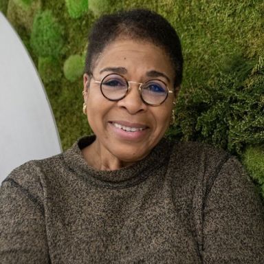 Smiling dark-skinned, female-presenting individual with short dark hair, with glasses, wearing a dark shirt in front of foliage