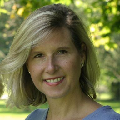 Smiling light-skinned, female-presenting individual with short blonde hair wearing a blue shirt on an outdoor backdrop