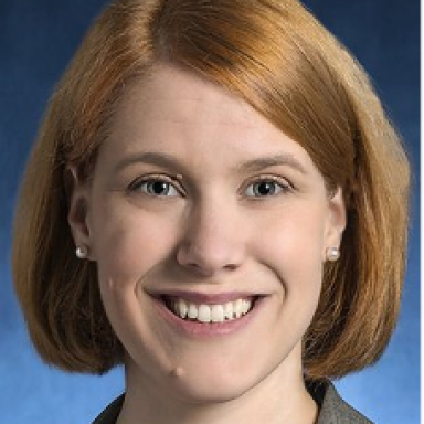 A smiling light skinned female with short red hair wearing a gray top against a blue background