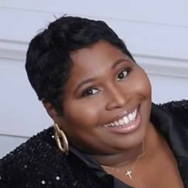 A smiling darker-skinned female with short black hair wearing a black jacket against a gray background