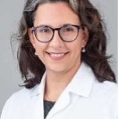 A smiling female with light skin and dark gray and brown hair wearing glasses and a lab coat against a light gray background