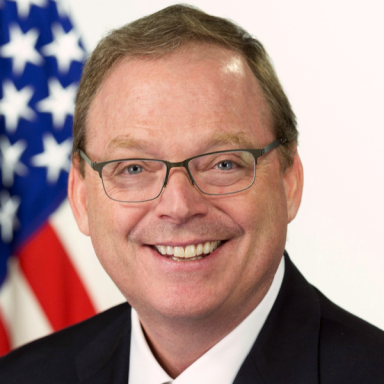 Smiling light-skinned, male-presenting individual with short light hair wearing a dark suit on a white backdrop, with an American flag.