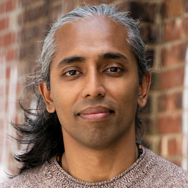 A darker-skinned male with long black and gray hair wearing a brown sweater against a brick wall background