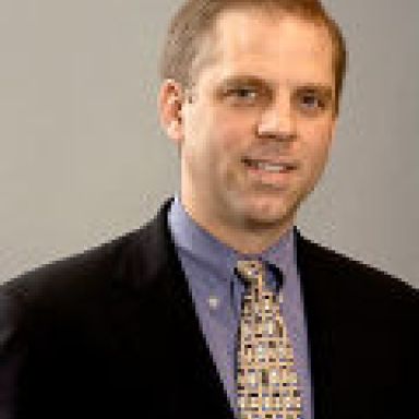 A smiling light-skinned male with light brown hair wearing a dark suit and tie against a gray background