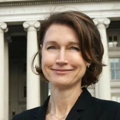 A smiling light-skinned female with short brown hair wearing a dark suit jacket against a building in the background