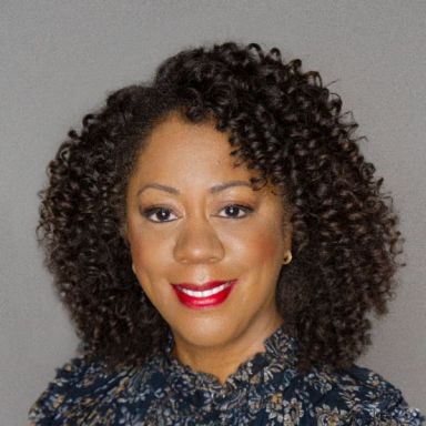 A smiling darker-skinned female with black curly hair wearing a dark patterned shirt against a gray background