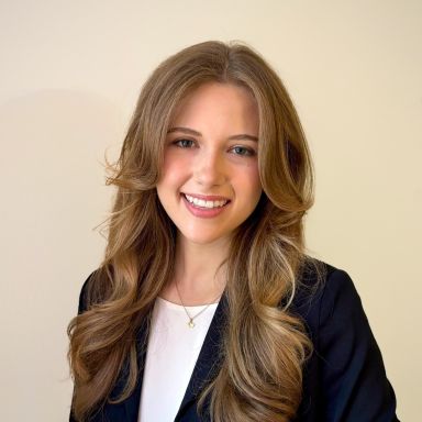 A smiling light-skinned female with long light brown hair wearing a dark suit jacket over a white shirt against a light background