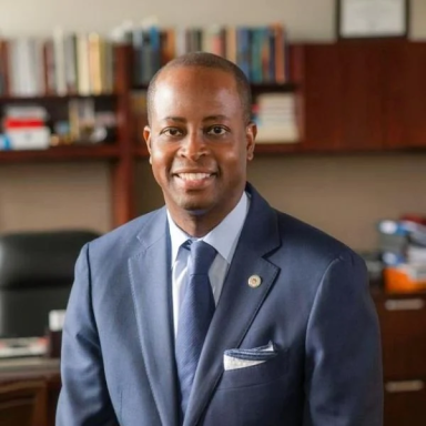 A smiling darker-skinned male wearing a blue suit and tie against a bookshelf in the background