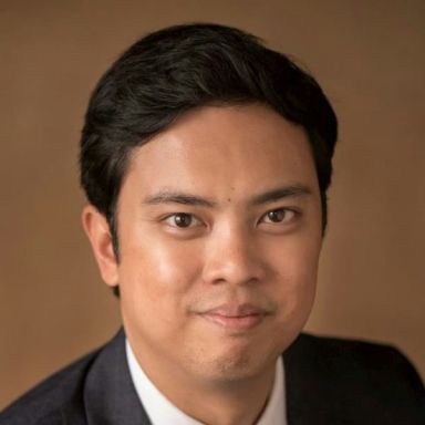 A smiling male of Asian heritage with black hair wearing a gray suit and tie against a grown background