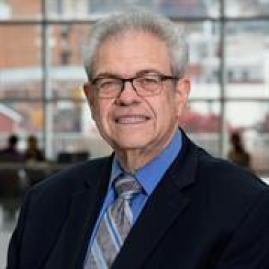 A smiling light-skinned male with gray hair wearing glasses and a dark suit and tie against an ambiguous background