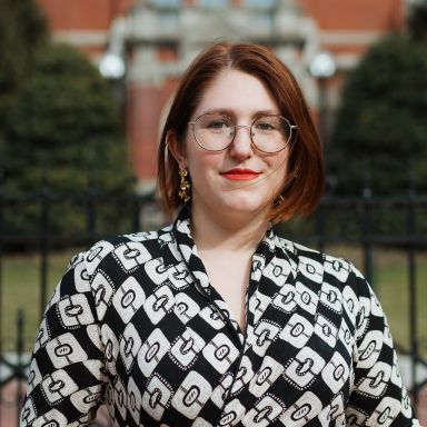 An woman with auburn hair and glasses in front of a building. 