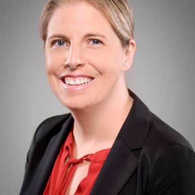 A smiling, light-haired woman wearing a red top and a black blazer against a gray background.