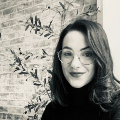 A smiling light-skinned female with dark brown hair wearing a black shirt and glasses against an ambiguous background